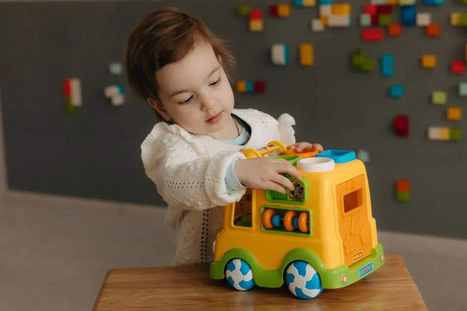 Un niño pequeño alcanza un autobús de juguete de colores sobre una mesa de madera en una sala de juegos luminosa