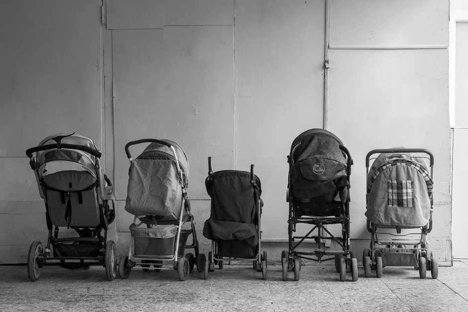 Five well-used baby strollers of different models lined up side by side against a wall in black and white, no people visible
