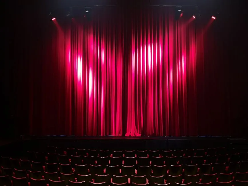Un telón de escenario rojo dramáticamente iluminado con focos sobre filas de butacas de teatro vacías en primer plano