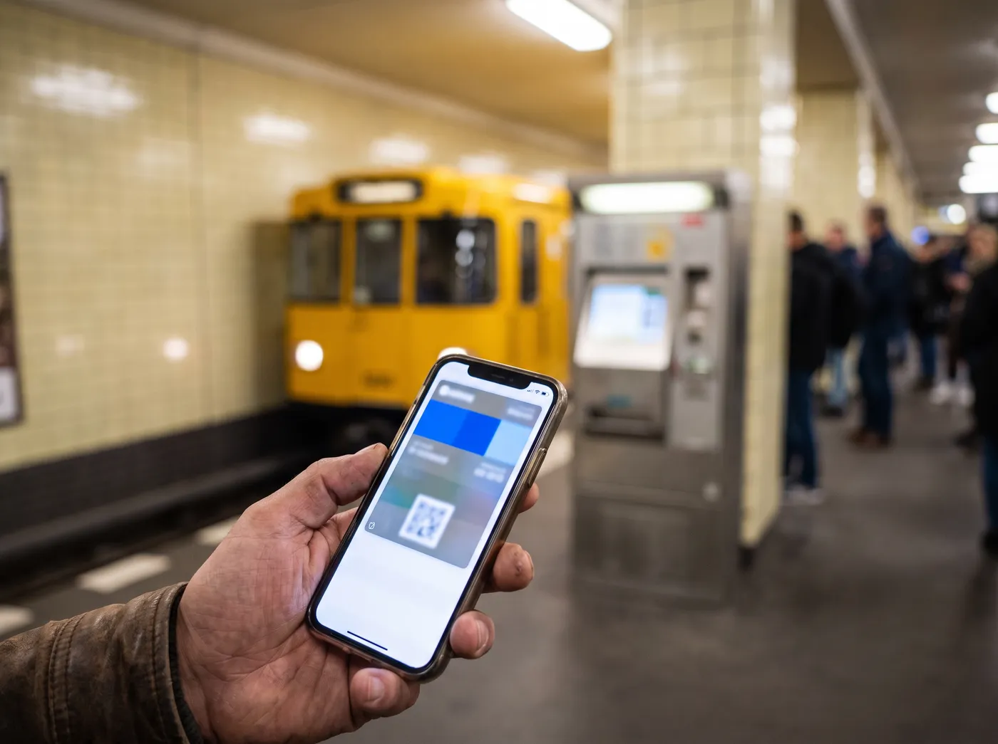 Eine Hand hält ein Smartphone mit einer Ticket-App auf einem Berliner U-Bahnsteig, im Hintergrund ein gelber Zug und ein Fahrscheinautomat