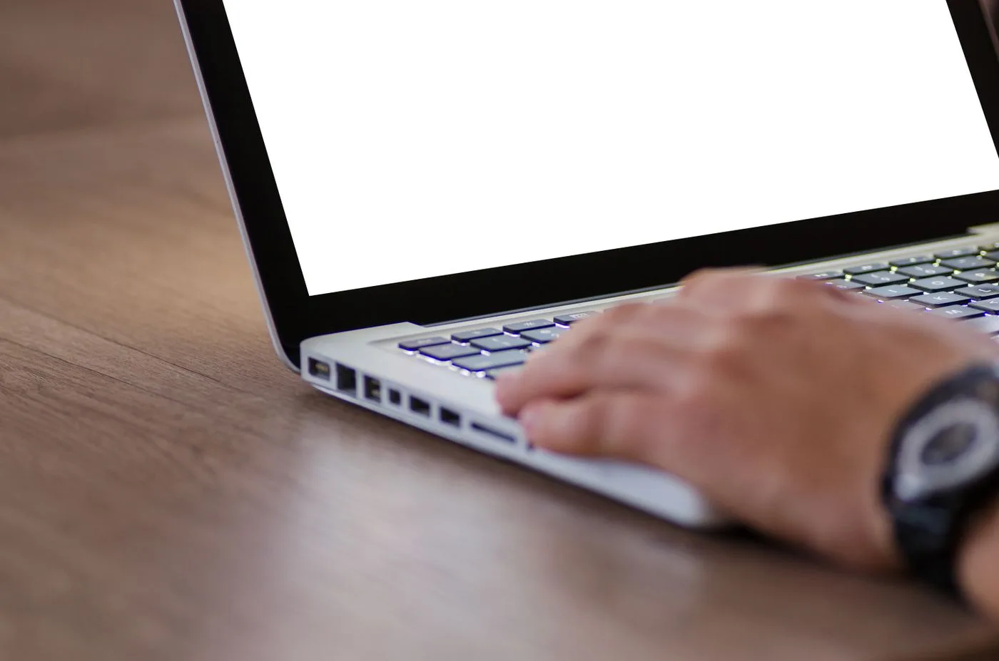 Primer plano de una mano escribiendo en el teclado de un portátil con una pantalla blanca en blanco, sobre un escritorio de madera