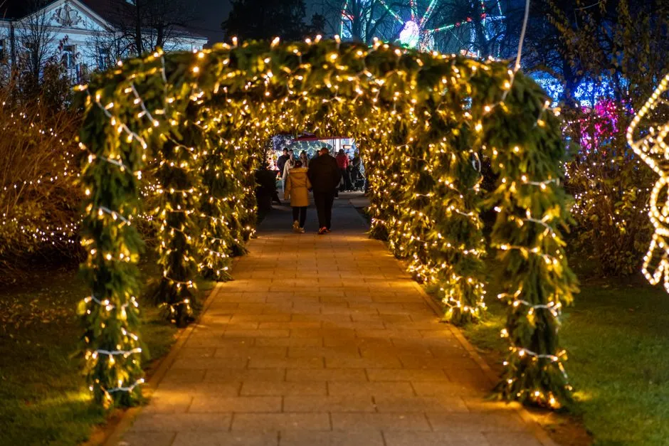 Ein Gartenweg bei Nacht unter einem mit warmen Lichterketten umwickelten Torbogen aus Tannenzweigen, im Hintergrund leuchten weitere beleuchtete Dekorationen entlang des Wegs