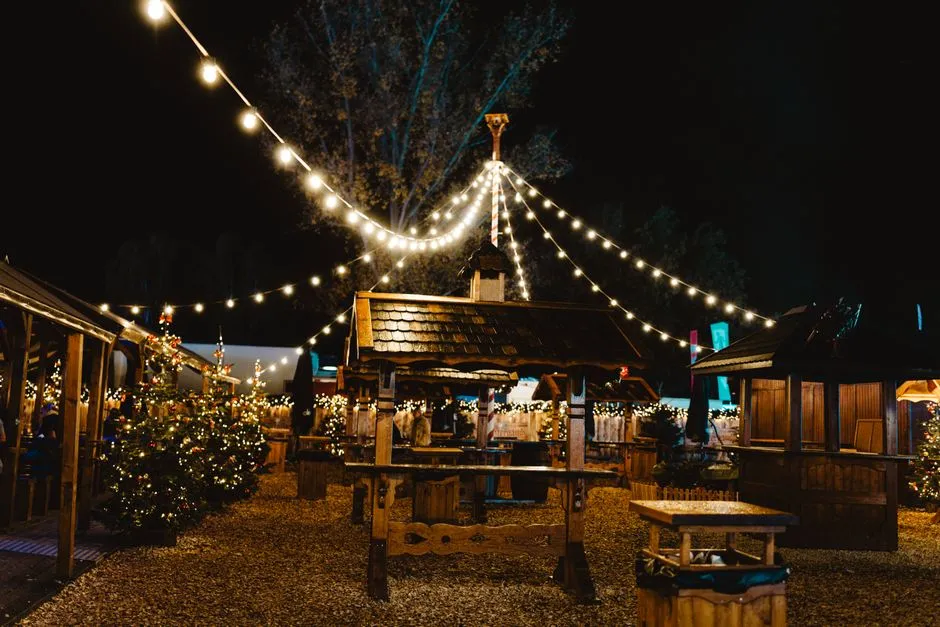Gece vakti sıcak ışıklı tellerle donatılmış, eğik ahşap kiremitli çatılara sahip ahşap Noel pazarı tezgahları, etrafta küçük süslenmiş Noel ağaçları