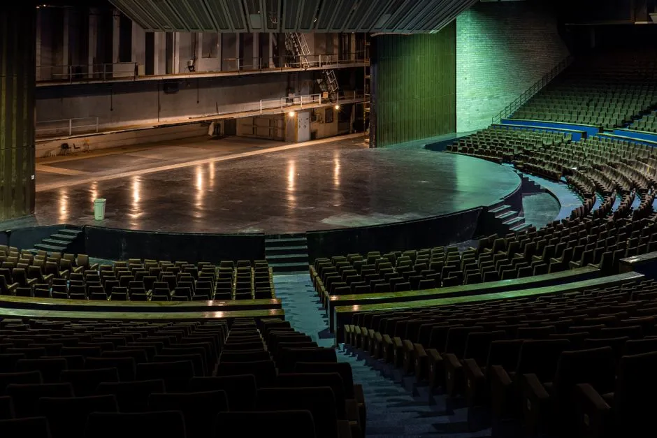 Leerer moderner Theatersaal von den hinteren Reihen aus gesehen, dunkelgrüne Sitze fallen zu einer runden, hell beleuchteten leeren Bühne ab, keine Personen sichtbar