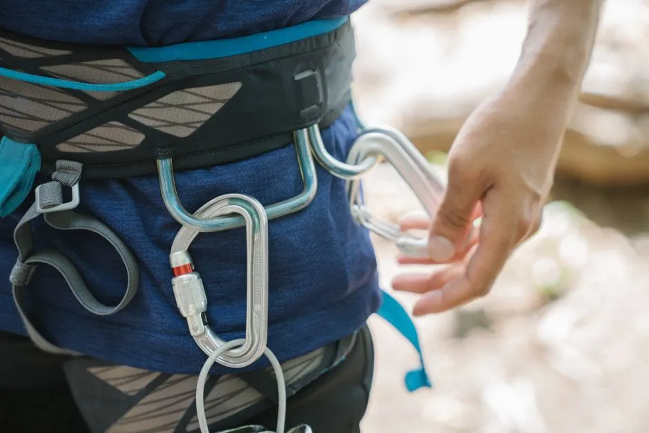 Nahaufnahme eines Klettergurts mit zwei eingehängten Karabinern, eine Hand greift nach einem dritten Karabiner, darunter ist ein Seil sichtbar