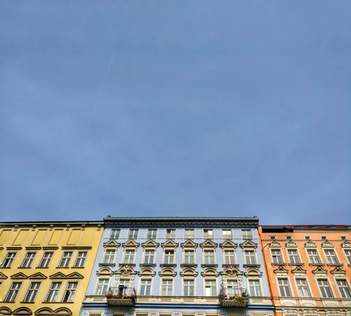 Drei unterschiedlich farbige historische Wohnhäuser, gelb, hellblau und orange, stehen nebeneinander unter einem klaren blauen Himmel, kein lesbarer Text oder Logos sichtbar