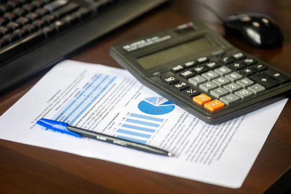 Ahşap bir masada, klavye ve fare yanında, çubuk ve pasta grafik içeren basılı bir finansal raporun üzerinde duran bir hesap makinesi