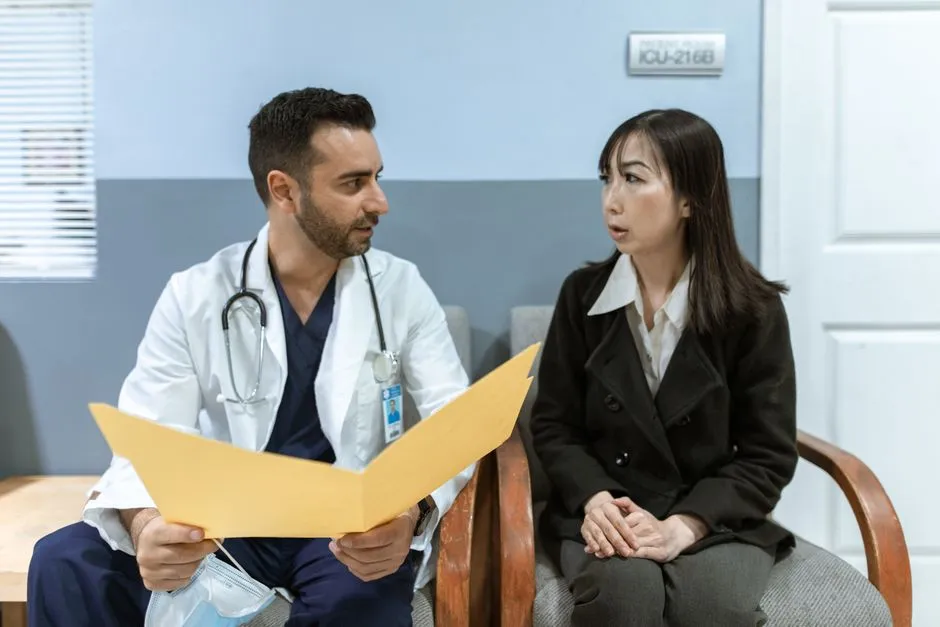 Ein Arzt im weißen Kittel sitzt mit einer Patientin in einem Wartebereich der Klinik und geht gemeinsam mit ihr eine Mappe mit Unterlagen durch
