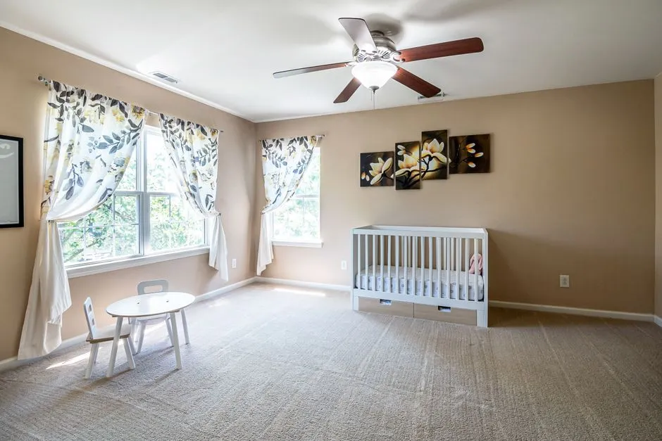 A white wooden baby crib in a bright, otherwise empty nursery room with a ceiling fan and curtains, no people or readable text visible