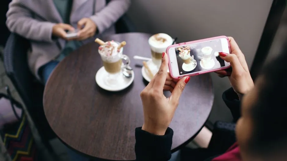 Zwei Personen an einem Cafétisch halten jeweils ein Smartphone hoch, um ihre Kaffeegetränke zu fotografieren, Hände und Telefone sichtbar, Gesichter außerhalb des Bildausschnitts