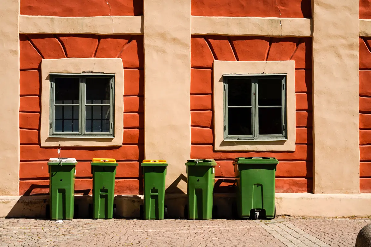 Fünf grüne Mülltonnen mit unterschiedlich farbigen Deckeln, aufgereiht vor einer sonnigen Hauswand, keine Gesichter, Logos oder lesbaren Beschilderungen sichtbar