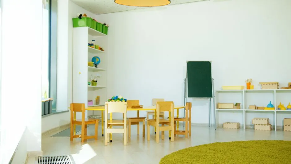 Una sala de juegos vacía de Kita con sillas y mesas pequeñas de madera, estanterías bajas llenas de juguetes y libros, un caballete de pizarra en blanco, sin personas ni texto legible