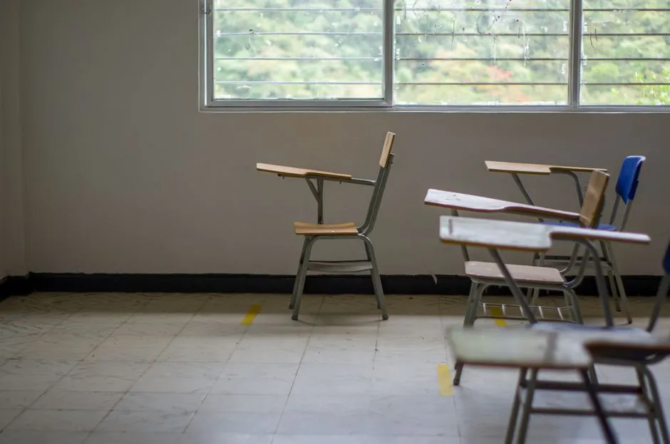 Ein leeres Klassenzimmer mit gestapelten Holzschulstühlen neben einem hellen Fenster, keine Personen oder lesbaren Texte sichtbar