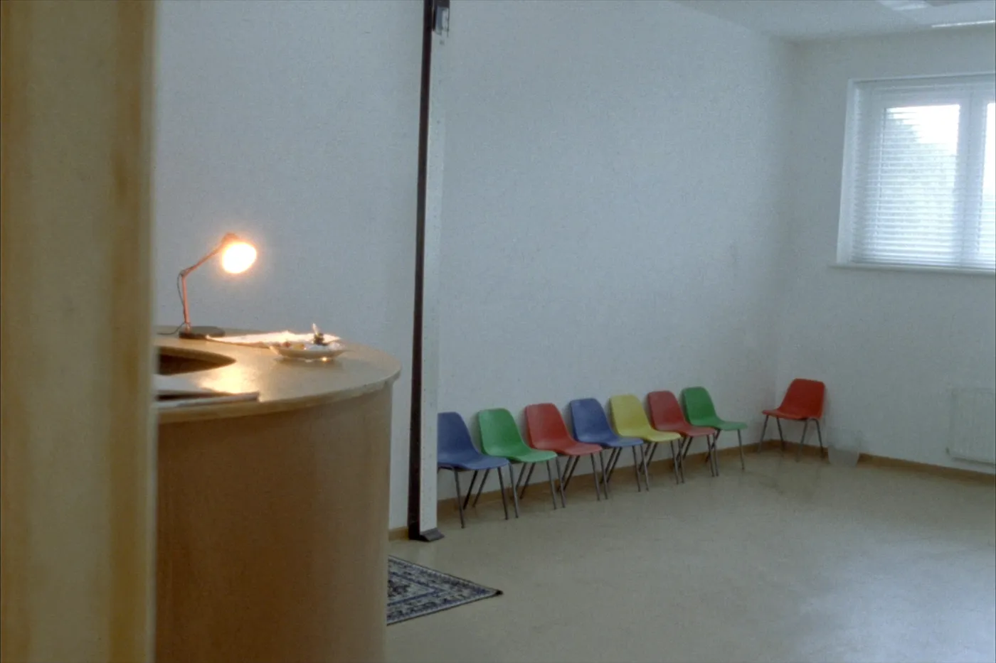 Ein ruhiger, leerer Kinderarztpraxis-Wartebereich mit einer Reihe kleiner bunter Stühle an einer schlichten Wand und einer warmen Empfangstisch-Lampe daneben