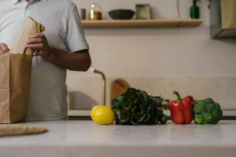 Oberkörper und Hände einer Person beim Auspacken einer braunen Papiertüte mit Lebensmitteln auf eine Küchenarbeitsplatte neben einer Zitrone, einem Kopfsalat, einer roten Paprika und Brokkoli, kein Gesicht sichtbar