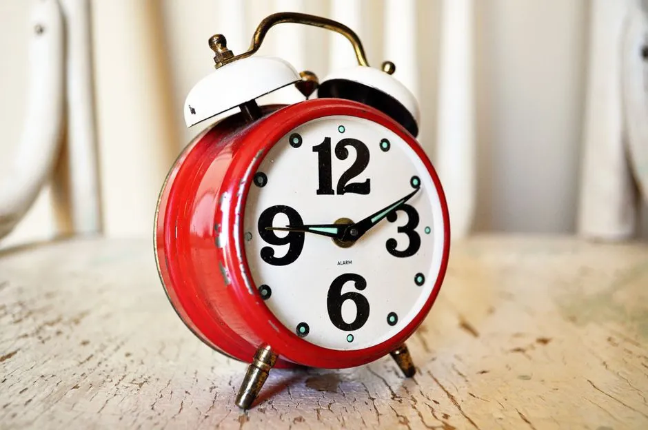 Un reloj despertador vintage rojo de doble campana con las manecillas señalando las dos en punto, sobre una superficie de madera, sin personas visibles