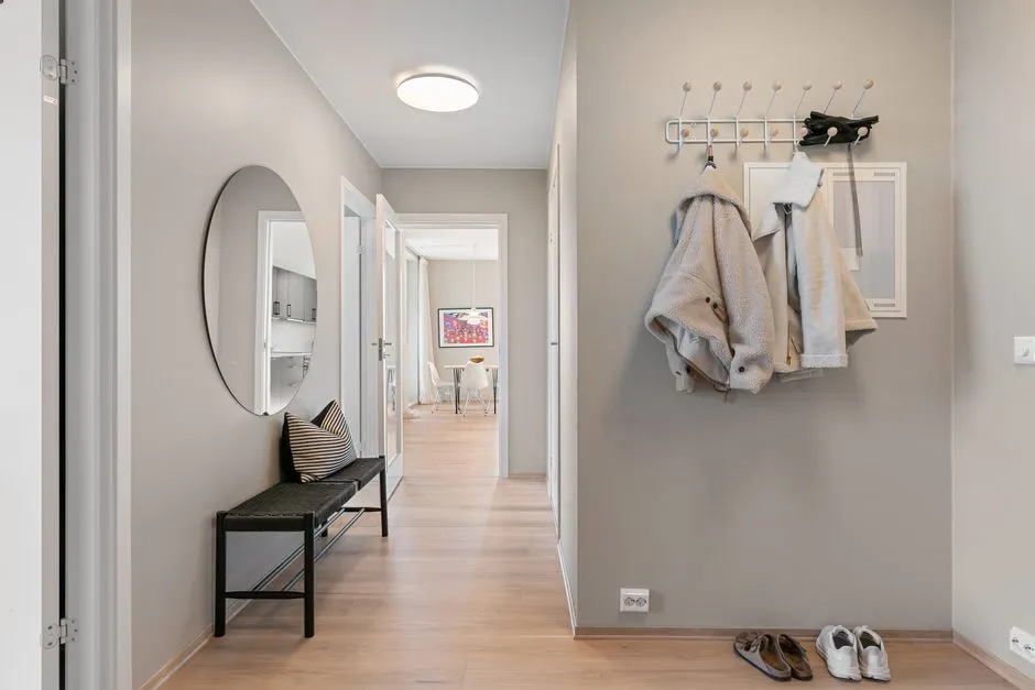 Ein minimalistischer europäischer Wohnungsflur mit einem runden Spiegel, einer dunklen Bank, an Wandhaken hängenden Jacken, und zwei Paar Schuhen, die ordentlich an der Wand neben dem Eingang stehen