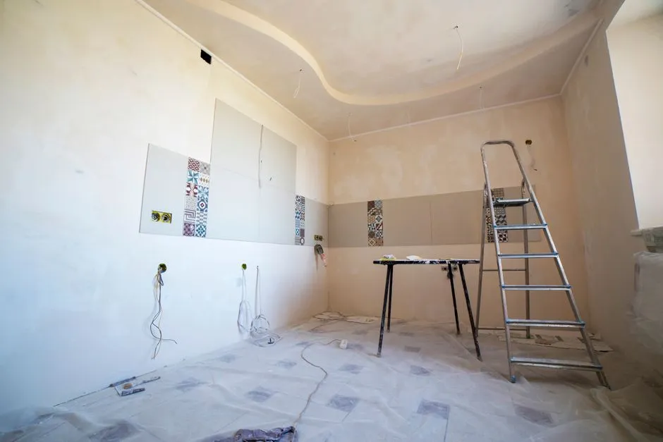 Habitación vacía en plena renovación con paredes enlucidas desnudas, muestras de baldosas apoyadas contra la pared, cables eléctricos a la vista, una escalera metálica, y una protección de suelo, sin armarios ni instalaciones de cocina