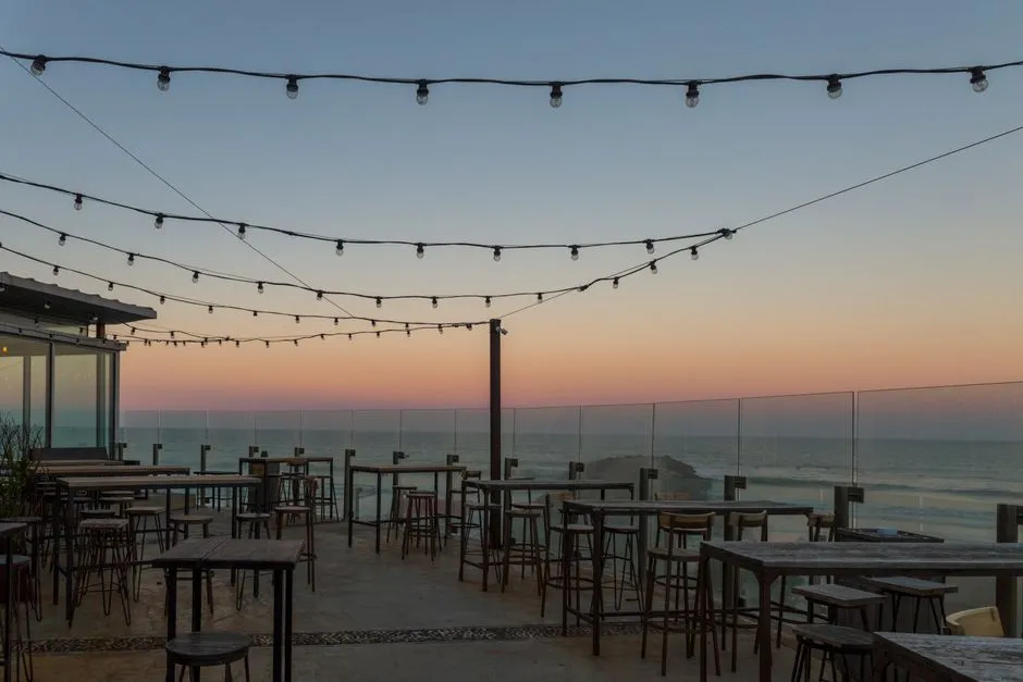 Eine leere Dachterrasse in der Abenddämmerung mit aufgehängten Lichterketten, leeren Bartischen und Hockern, und einem Glasgeländer mit Blick auf Meer und Himmel bei Sonnenuntergang