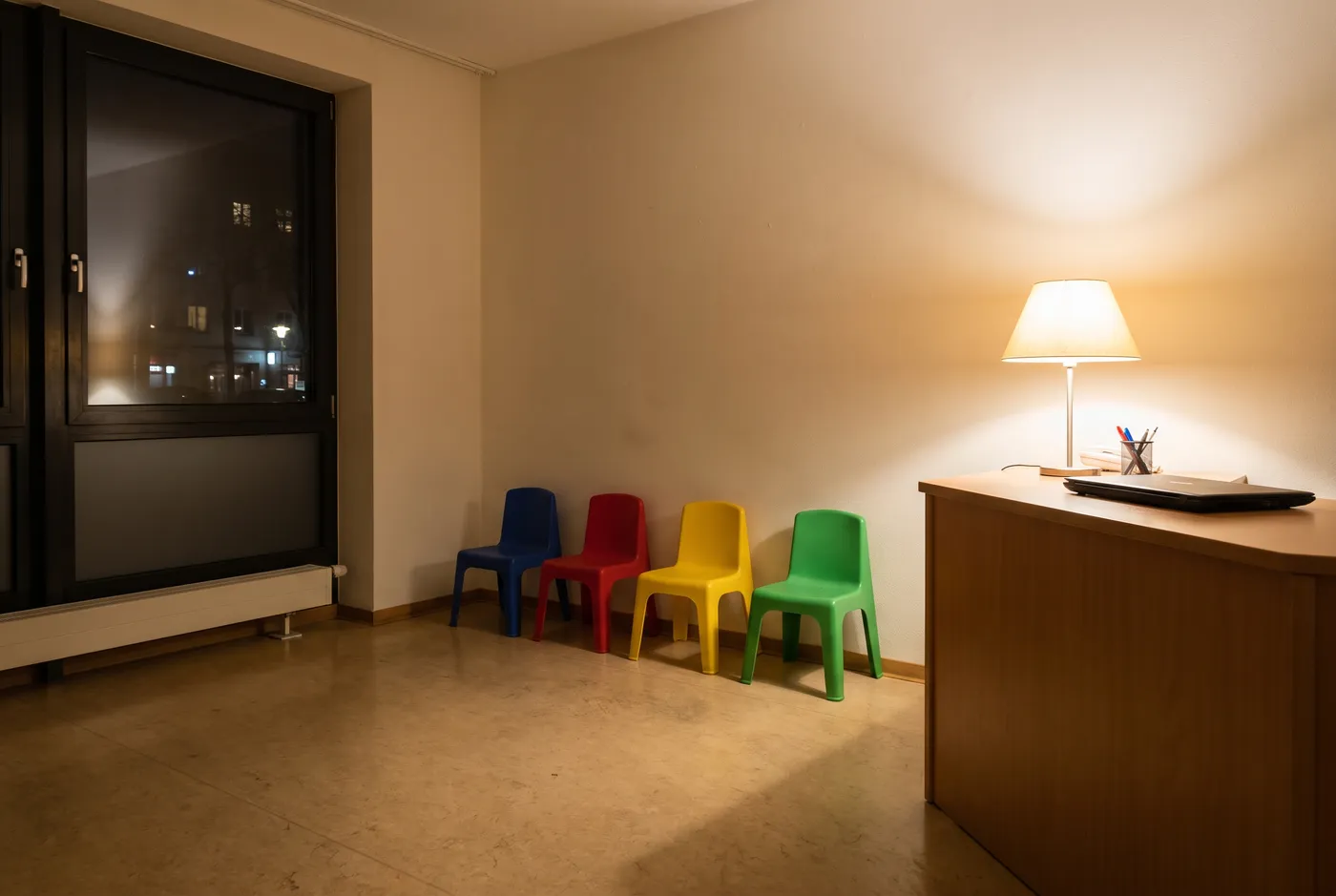 Ein leeres kleines Arztpraxis-Wartezimmer bei Nacht, vier bunte Kinderstühle an einer Wand, eine warme Tischlampe am Empfangstresen beleuchtet, ein dunkles Fenster mit fernen Stadtlichtern
