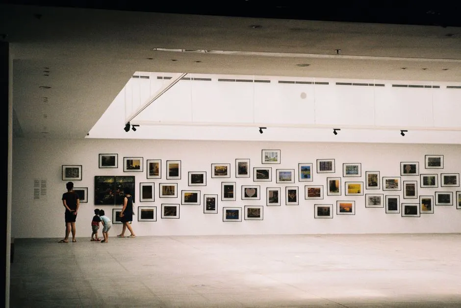 Ein Elternteil und zwei Kinder von hinten gesehen, sie stehen mit Abstand und betrachten eine Wand voller kleiner gerahmter Fotografien in einer hellen, minimalistischen Kunstgalerie