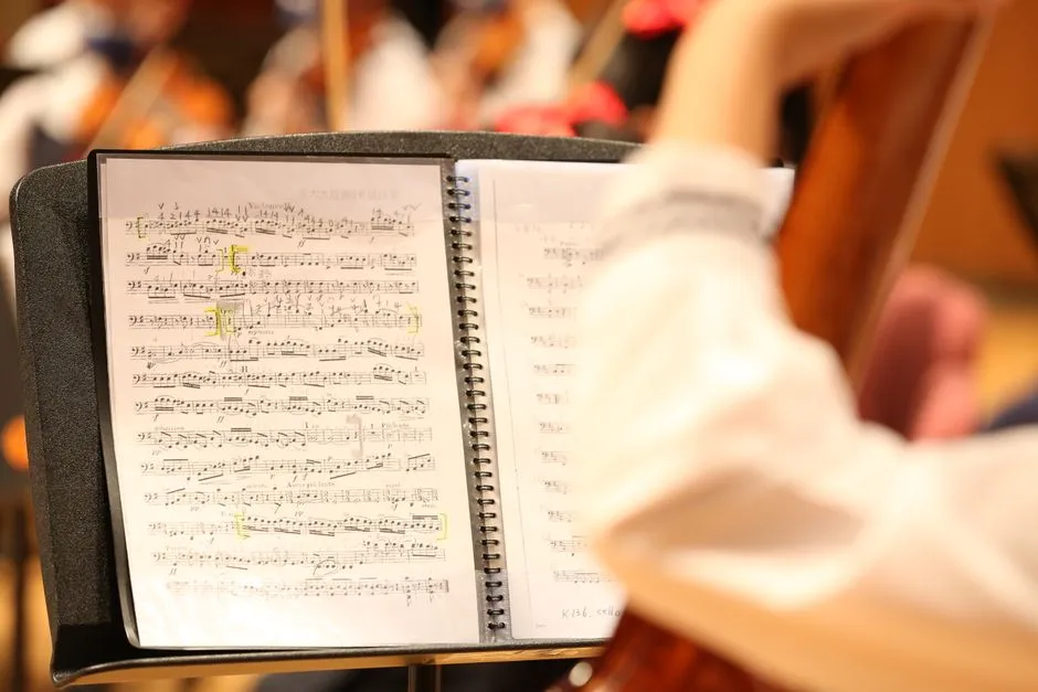 Nahaufnahme eines aufgeschlagenen Notenordners auf einem Orchesternotenständer während einer Probe, der Arm einer Musikerin oder eines Musikers greift nach der Seite, verschwommen im Hintergrund sind Streicherinnen und Streicher sowie Instrumente zu erkennen, keine Gesichter sichtbar