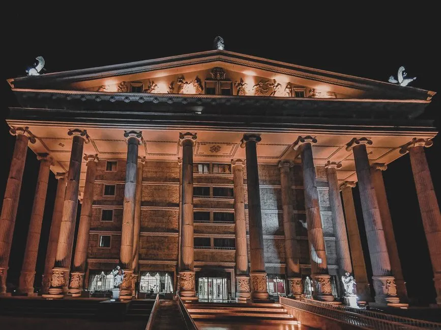 Un gran edificio de museo con altas columnas clásicas y un frontón triangular ornamentado, iluminado en luz naranja cálida contra un cielo nocturno completamente oscuro