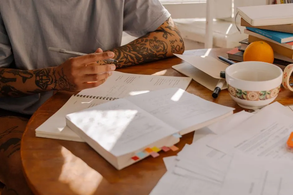 Ein tätowierter Unterarm ruht auf einem Holztisch voller offener Notizhefte, loser Prüfungsblätter, eines Stifts und einer Tasse Tee, Handschrift zu klein zum Lesen, kein Gesicht sichtbar