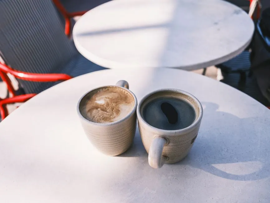 Dos tazas de café de cerámica una junto a la otra sobre una mesa redonda de cafetería al aire libre bajo luz solar cálida, una cubierta con espuma de leche, sillas vacías visibles en una mesa vecina