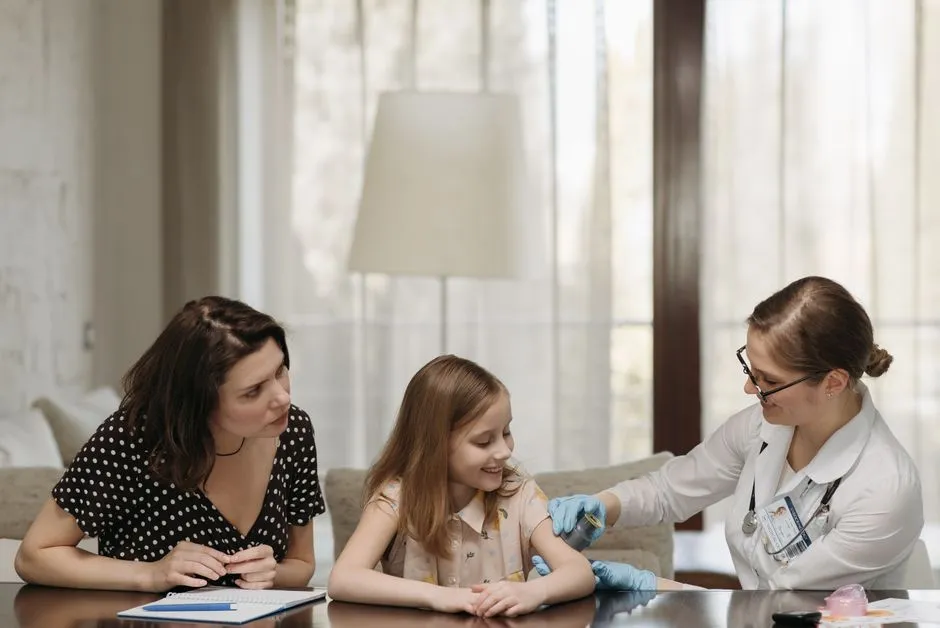 A doctor in a white coat administering a vaccination to a smiling young girl at a table while her mother watches closely