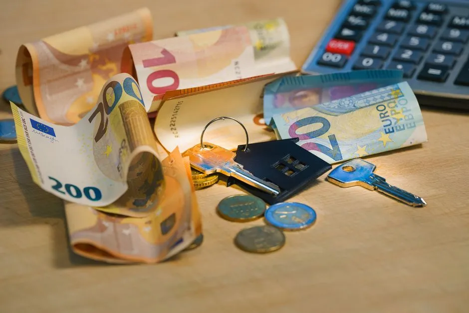 Euro banknotes, coins, a house-shaped keychain, and a calculator on a wooden table, representing the cash sum a rental deposit ties up