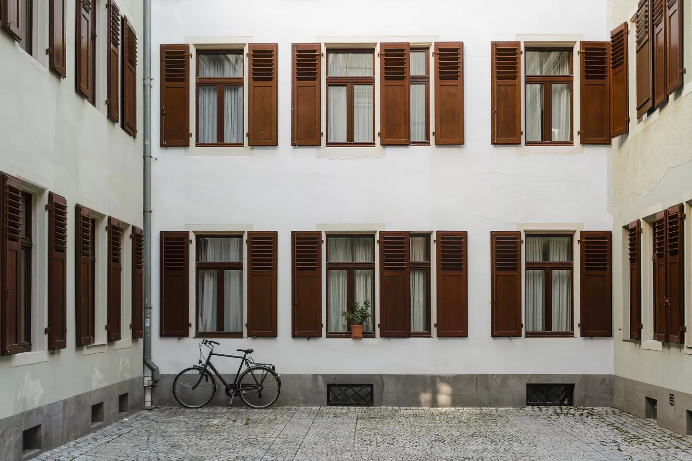 Un patio tranquilo de Altbau de Berlín al mediodía, contraventanas de madera cerradas, una bicicleta apoyada en la pared, una planta en maceta en el alféizar de una ventana, adoquines vacíos