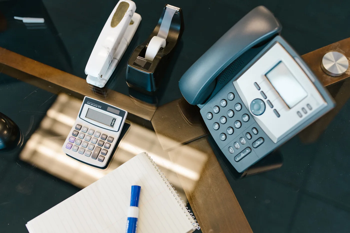 Ein von oben fotografierter Bürotisch mit Telefon, Taschenrechner, Spiralnotizblock, Stift und Klebebandabroller