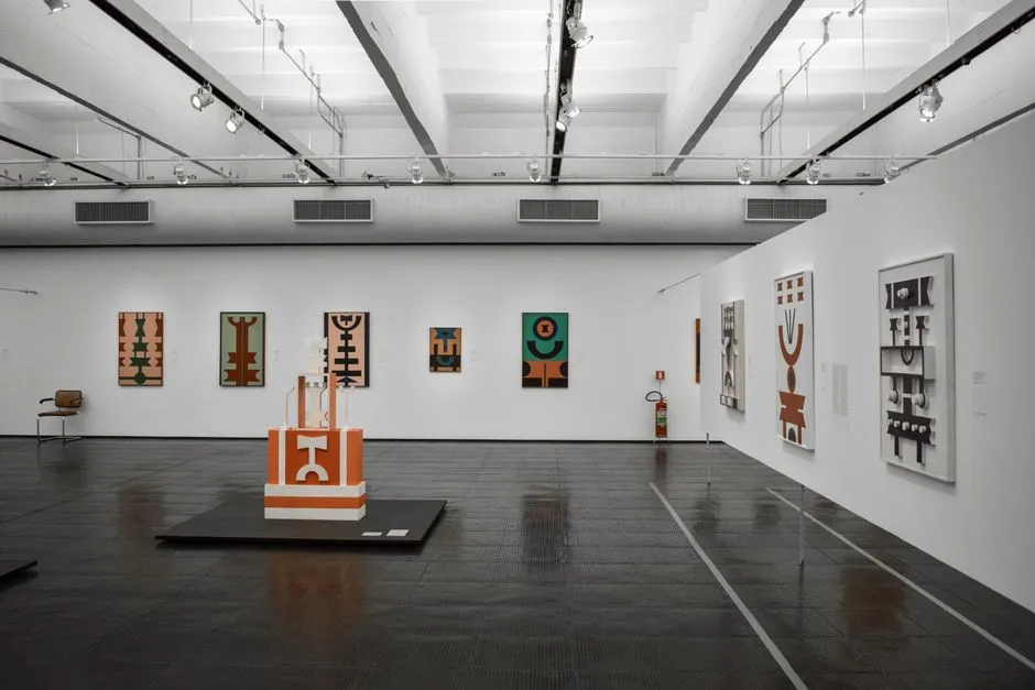 Una sala de galería blanca minimalista y vacía con una fila de pequeños grabados abstractos en una pared y una escultura geométrica naranja mostrada sobre un pedestal bajo en el centro
