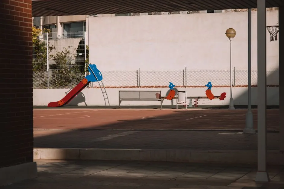 Ein leerer Schulhof-Spielplatz mit einer roten Rutsche, Federwippen, einer Bank und einem Basketballkorb, umzäunt vor einer schlichten Wand, keine Personen anwesend
