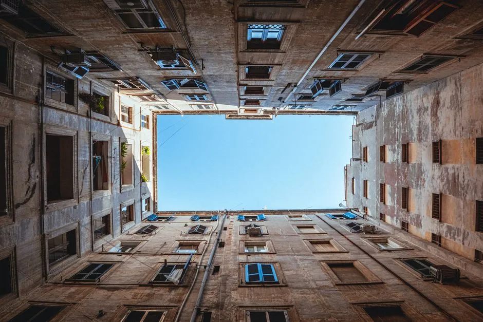 Mirando directamente hacia arriba desde el suelo dentro de un patio encerrado de estilo Berlín, rodeado en los cuatro lados por las fachadas con ventanas de edificios de apartamentos antiguos, cielo abierto visible en el centro, sin personas visibles