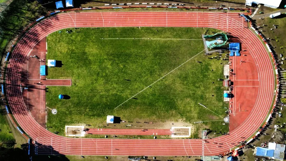 Luftaufnahme einer roten und weißen Leichtathletik-Laufbahn rund um ein grünes Stadioninnenfeld, markierte Bahnlinien sichtbar, keine Gesichter erkennbar