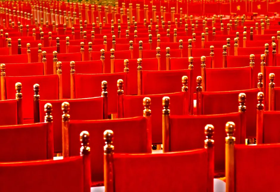 Reihen leerer roter Samtstühle mit Messingknäufen in einem Theater- oder Konzertsaal, keine Personen sichtbar