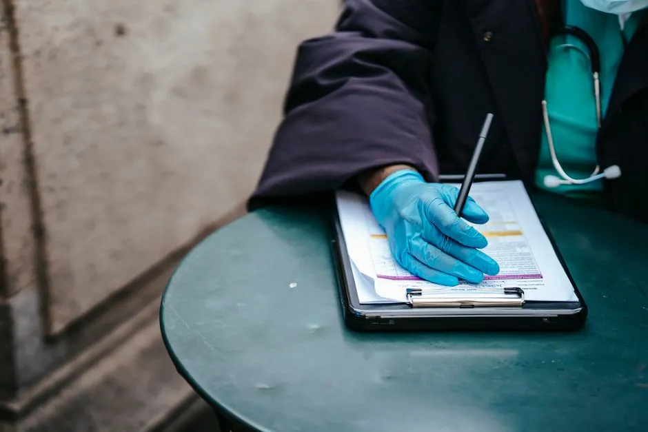 A gloved hand holding a pen over a clipboard of medical paperwork, with a stethoscope resting nearby on an outdoor table