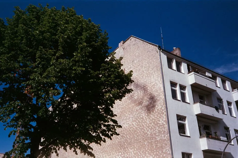 Un edificio residencial de apartamentos en Berlín visto desde abajo, con fachada de estuco pálido, balcones, y un gran árbol en primer plano
