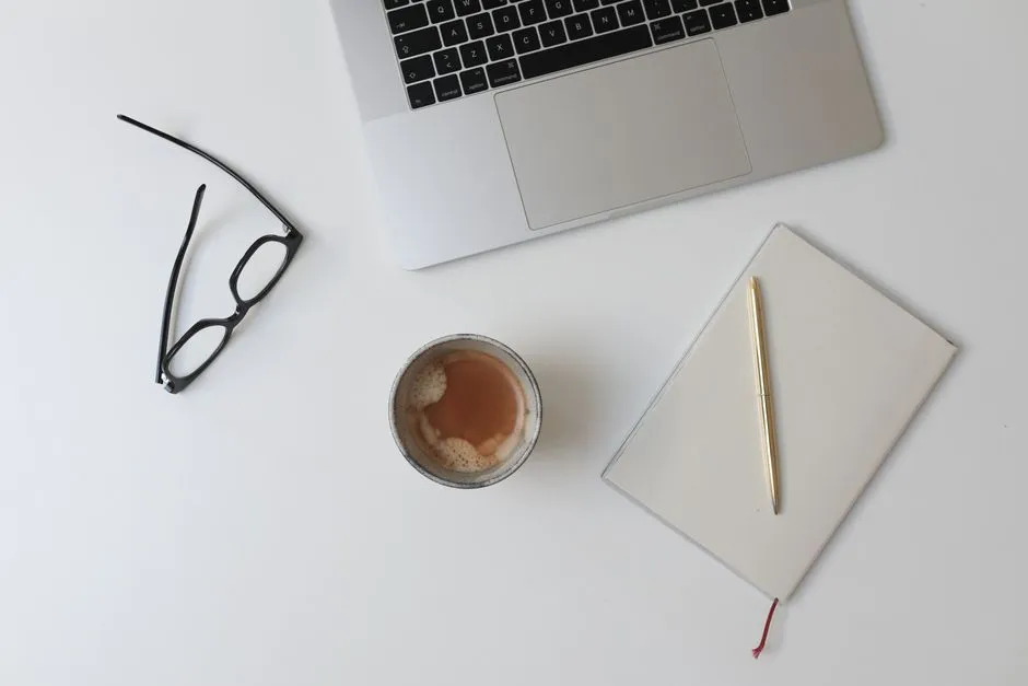 Ein minimalistischer weißer Schreibtisch von oben gesehen, mit Lesebrille, einer Tasse Kaffee, einem geschlossenen Notizbuch, einem Stift und dem Rand einer Laptop-Tastatur