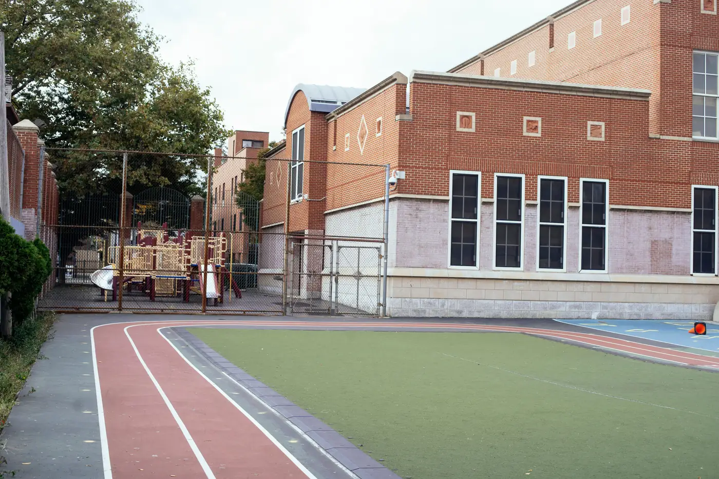 Ein zeitgenössisches Backsteinschulgebäude mit eingezäunter Laufbahn und Rasensportplatz, Spielgeräte hinter einem Maschendrahtzaun sichtbar, keine Menschen oder lesbaren Texte oder Logos