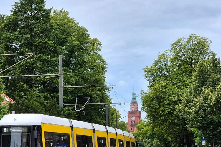 Eine gelbe BVG-Straßenbahn fährt unter Oberleitungen an Bäumen vorbei, im Hintergrund ein roter Backstein-Uhrturm auf einer Berliner Straße sichtbar