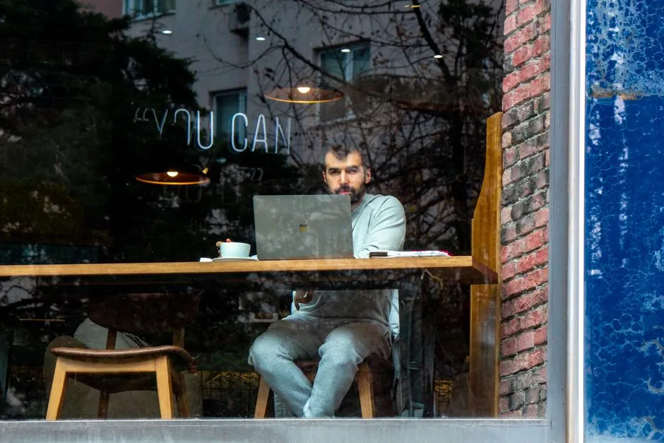Ein Mann arbeitet an einem Laptop an einem Fenstertisch in einem Café, von außen durch das Glas gesehen, eine Kaffeetasse und ein Notizbuch neben ihm, Bäume und ein Gebäude spiegeln sich im Fenster