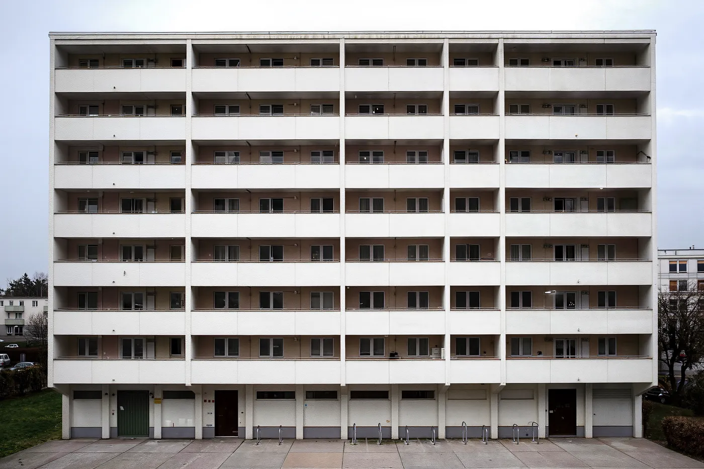 A wide, plain 1970s-style Berlin apartment block with rows of identical balconies and bicycle racks along the ground floor, no people or readable signage visible