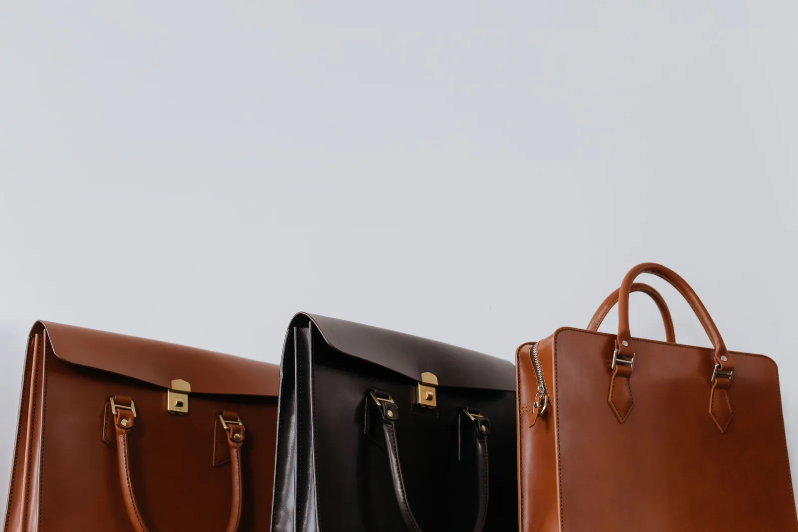 Drei Ledertaschen in Braun, Schwarz und Beige, nebeneinander vor einer schlichten hellgrauen Wand angeordnet, keine Gesichter oder lesbarer Text sichtbar