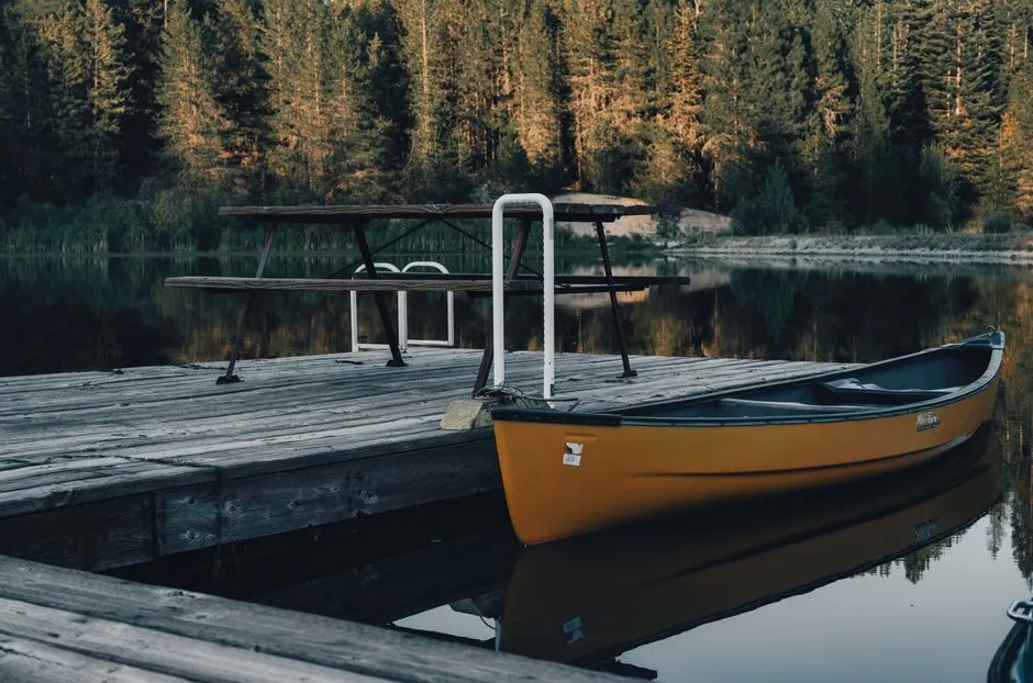 Ein einzelnes Holzkanu, festgemacht an einem ruhigen Seesteg, ein Paddel liegt in der Nähe, Kiefernwald spiegelt sich im stillen Wasser, keine Menschen sichtbar