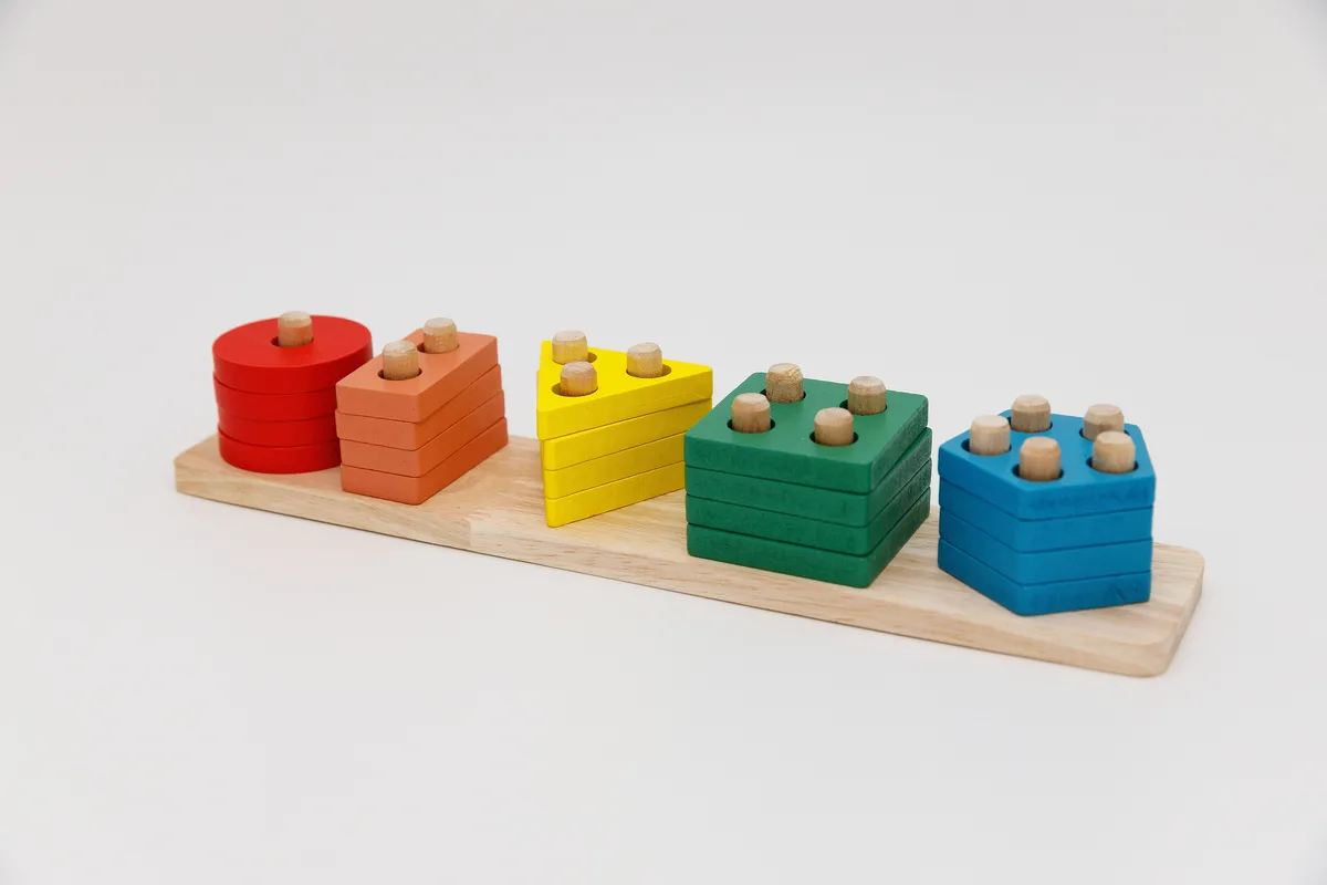 Eine Reihe bunter, nach Größe gestaffelter Holz-Stapelspielzeuge, von einem kleinen roten Zylinder bis zu einer größeren blauen sechseckigen Form, auf einem schlichten Holzbrett
