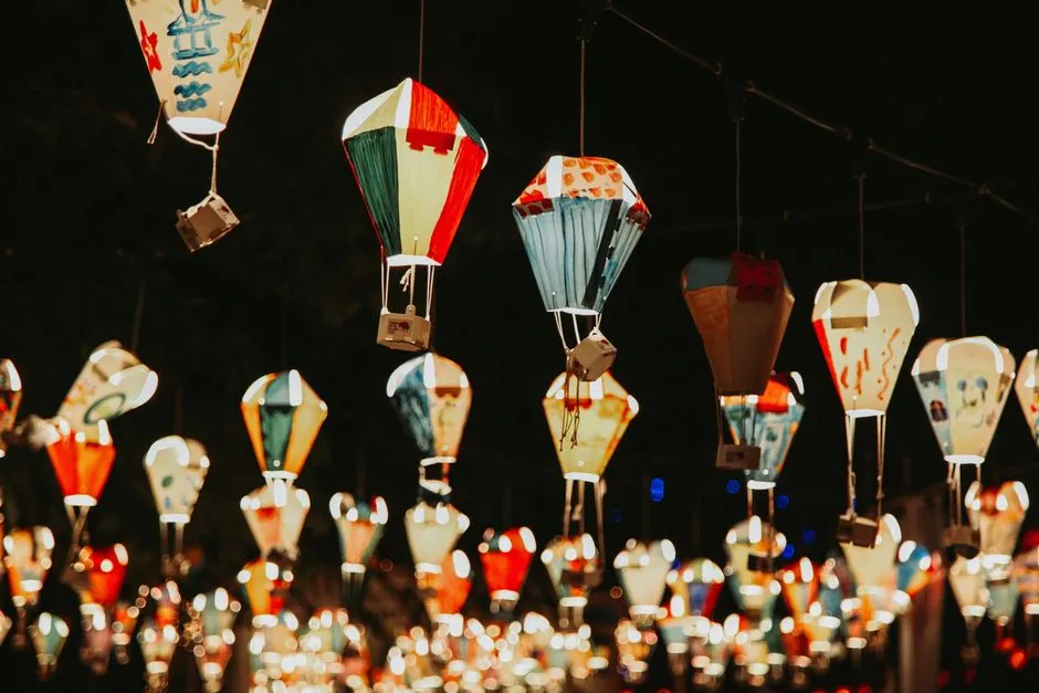 Farolillos de papel coloridos pintados a mano con forma de pequeños globos aerostáticos, colgados en líneas y brillando de noche, decorados con dibujos infantiles simples