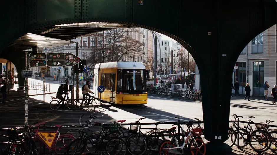 Un tranvía amarillo de Berlín pasando bajo un puente ferroviario en una calle soleada, con filas de bicicletas aparcadas en primer plano y dos ciclistas montando junto al tranvía